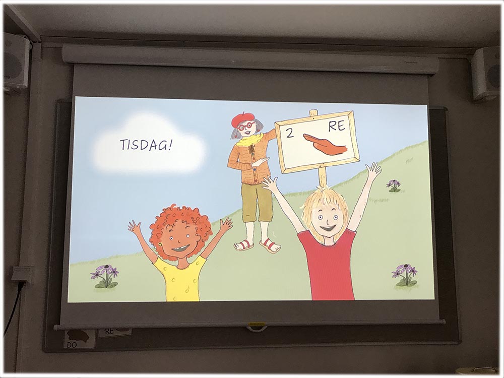 En projektorduk där en tecknad bild från E-boken Tonförsbacken visas. Två glada barn med armarna uppsträckta i förgrunden och i bakgrunden dam i röd basker som står vid en skylt i en grön backe. På skylten står det RE (som i do - re -mi) och på ett moln mot den blå himlen står det tisdag.