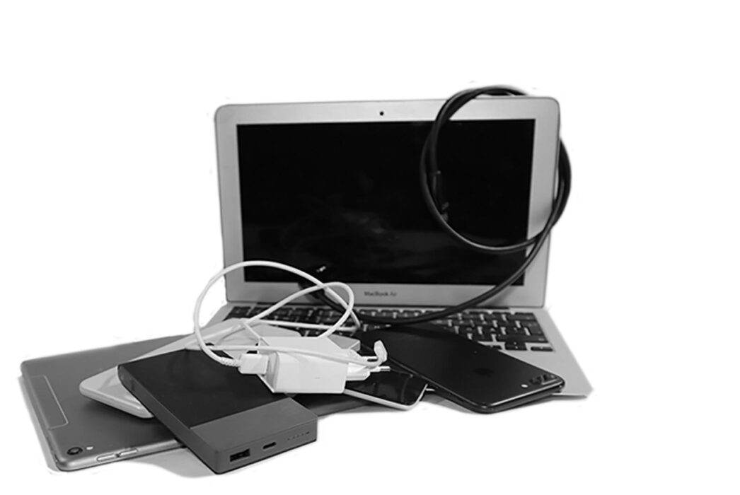 Svartvit bild med en uppslagen laptop, en massa sladdar, iPad, mobil, laddare i en enda röra!